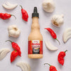 A bottle of Frank's RedHot Creamy Garlic Sauce is surrounded by garlic bulbs, cloves, and red chili peppers.