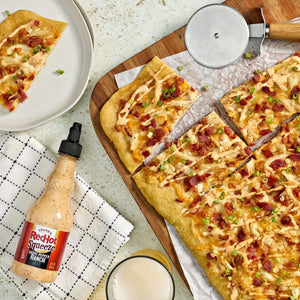 A flatbread pizza topped with bacon and sauce, alongside a bottle of Burklees sauce and a pizza cutter.