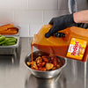 A gloved hand pours Frank's RedHot Wings Spicy Maple Sauce onto fried chicken pieces in a metal bowl, with celery and carrot sticks in metal containers in the background.