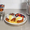 A plate features two waffles topped with a savory topping and poached eggs covered in hollandaise sauce, accompanied by a side of fresh strawberries and blueberries, and a small dish of sauce.