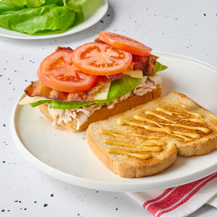 An open-faced sandwich with turkey, bacon, tomato, lettuce, cheese, and mustard on toasted bread is displayed on a white plate.