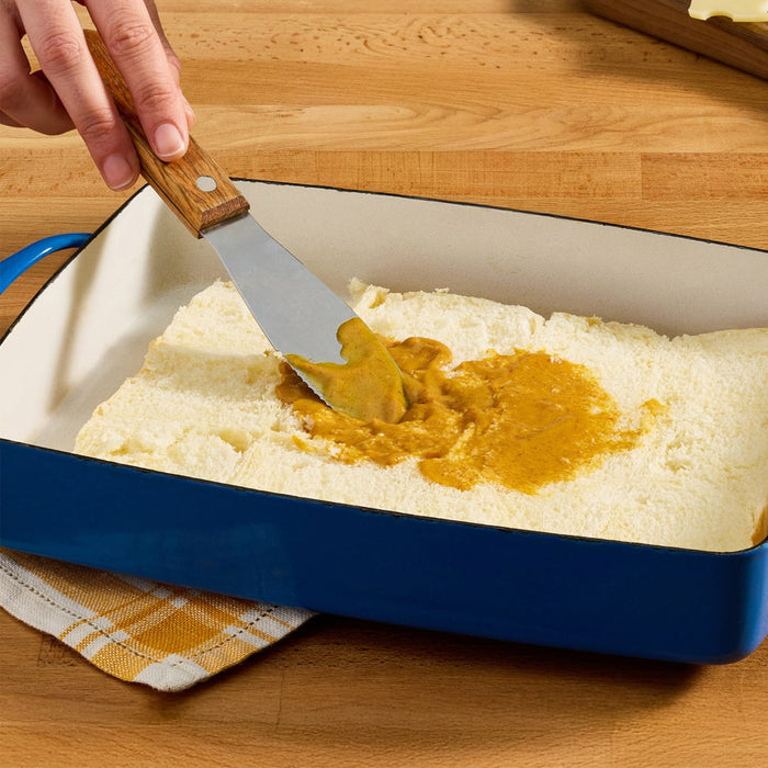A person spreads a brown sauce over a layer of white cake in a blue baking dish.