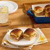 A plate of delicious ham and cheese slider sandwiches is presented alongside a refreshing drink and a baking dish full of more sliders.