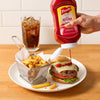 A hand squeezes a bottle of FrenchS Tomato Ketchup, 20 Oz onto a cheeseburger served with a side of fries and a cold soda.