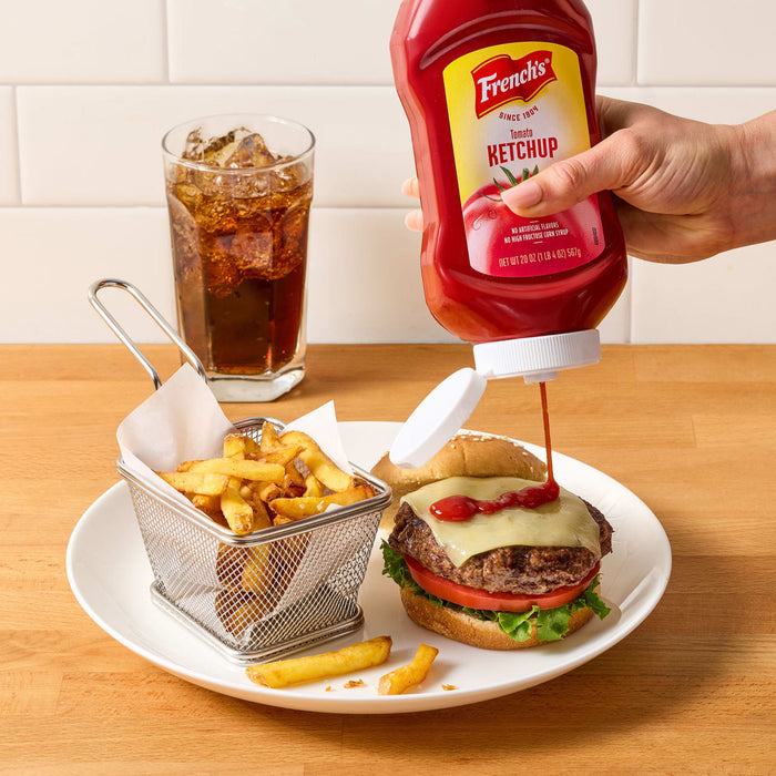 A hand squeezes a bottle of FrenchS Tomato Ketchup, 20 Oz onto a cheeseburger served with a side of fries and a cold soda.
