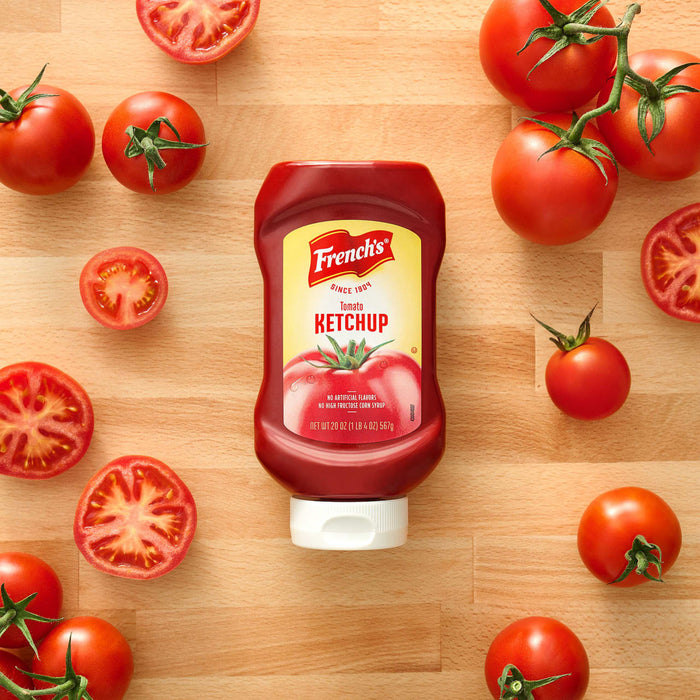 A bottle of FrenchS Tomato Ketchup, 20 Oz is centered on a wooden surface surrounded by fresh whole and sliced red tomatoes.