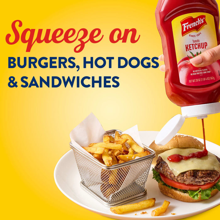 A hand squeezes French S Tomato Ketchup, 20 Oz onto a cheeseburger served with a side of golden fries in a wire basket against a bright yellow background.