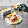 A gloved hand ladles a creamy portion of Cattlemen'S Alabama White Bbq Sauce, 0.5 Gal onto a freshly made roast beef sandwich, served with a pickle and a side of french fries.