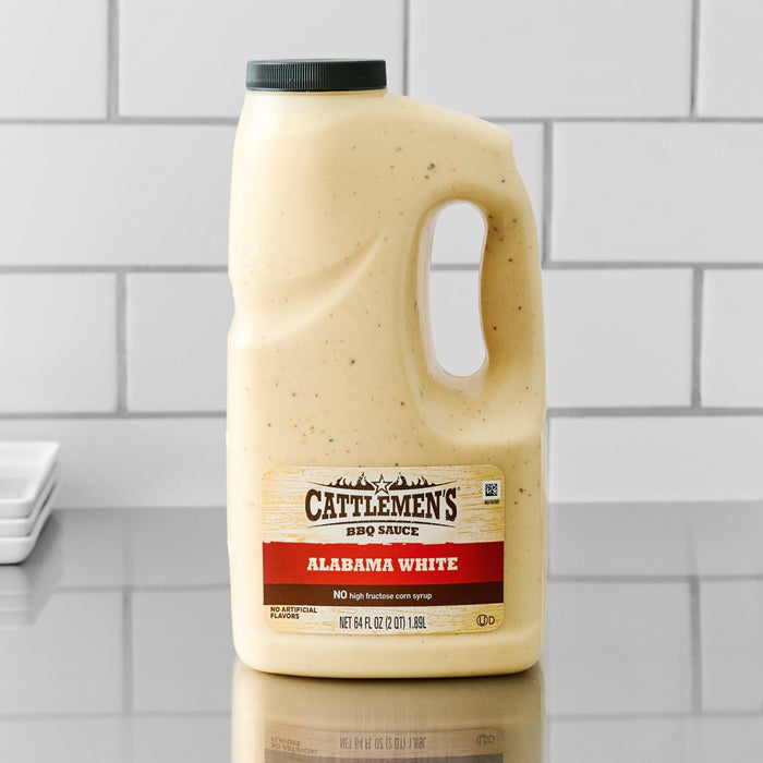 A large, half-gallon jug of Cattlemen'S Alabama White Bbq Sauce, 0.5 Gal sits on a stainless steel countertop, ready for use in a professional kitchen.