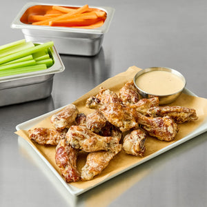 A platter of chicken wings is generously coated and served with a side of Cattlemen'S Alabama White Bbq Sauce, 0.5 Gal, accompanied by fresh celery and carrot sticks.