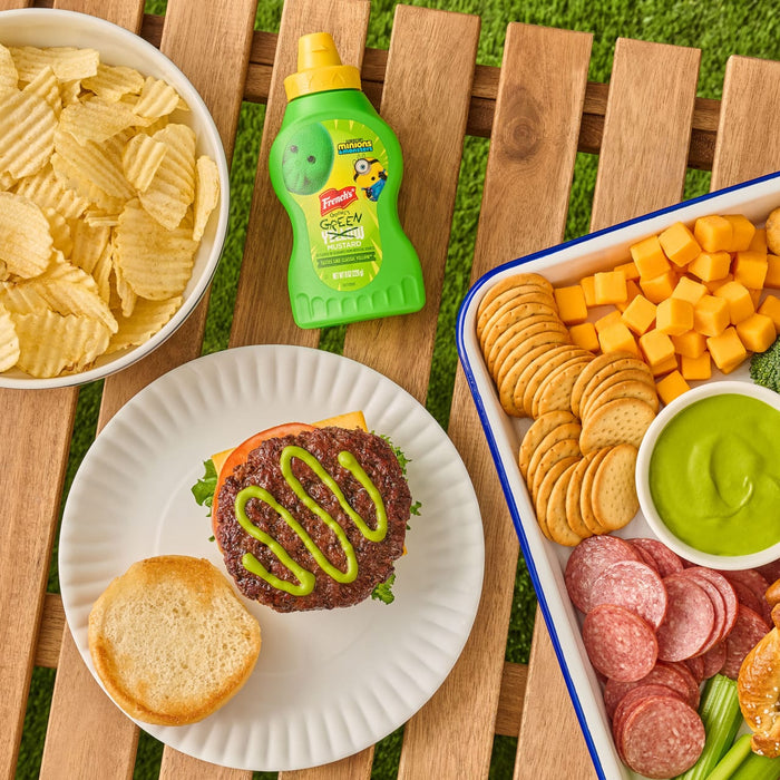 A bottle of French'S Goomi'S Green Mustard, 8.0 Oz, is shown on a picnic table next to a hamburger topped with the green mustard, a bowl of chips, and a snack tray.