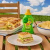 A bottle of French'S Goomi'S Green Mustard, 8.0 Oz, is displayed on a picnic table next to a plate of ham and cheese sliders made with the green mustard.