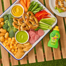 A colorful snack board perfect for a picnic features pretzels, cheese, and vegetables arranged around a bowl of green dip, with a bottle of French'S Goomi'S Green Mustard, 8.0 Oz, sitting on the table nearby.