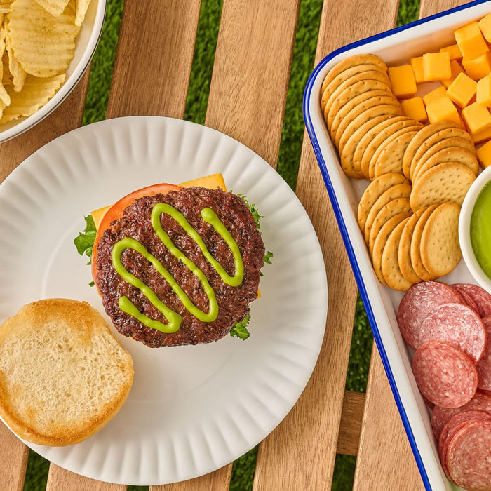 An open-faced hamburger on a paper plate is drizzled with French'S Goomi'S Green Mustard, 8.0 Oz, and served at a picnic with chips and a snack tray.