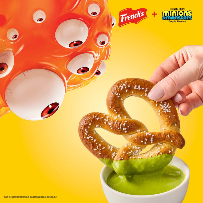 In a promotional illustration for French's and Illumination's Minions &amp; Monsters, a hand dips a pretzel into a bowl of French'S Goomi'S Green Mustard, 8.0 Oz, as an orange, multi-eyed monster looks on.