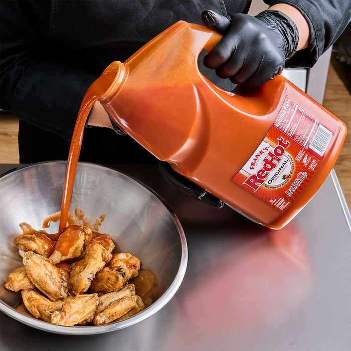 A hand in a black glove pours Frank's RedHot Original Cayenne Pepper Sauce over chicken wings in a bowl.