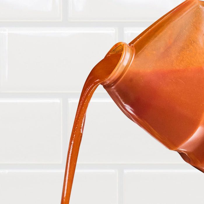 A bottle of orange sauce is pouring out onto a white tiled surface.