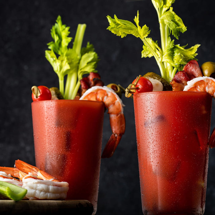 Two glasses of bloody mary garnished with shrimp, celery, bacon, olives, and a cherry.
