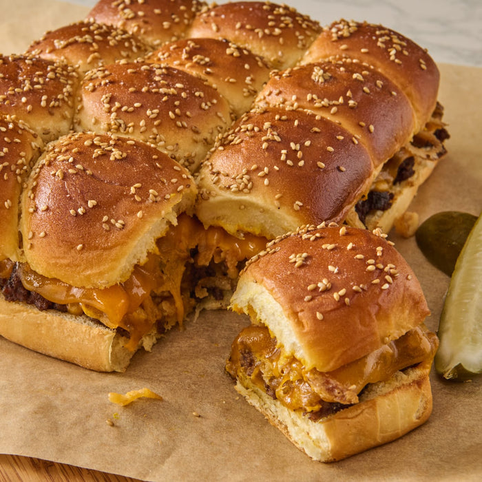 ['A close-up of a plate of mini cheeseburgers with sesame seed buns and pickles.']