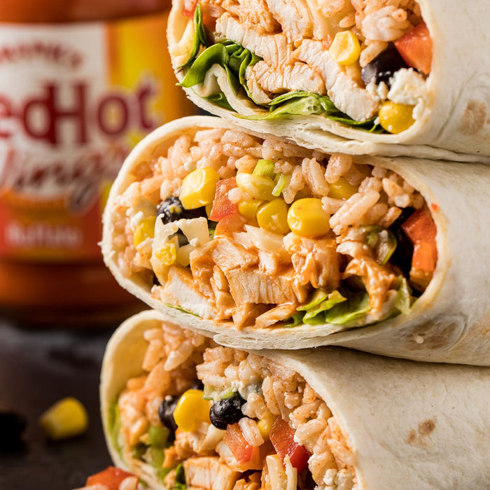 ['A stack of three burritos with chicken, rice, corn, black beans, lettuce, and red hot sauce.']