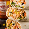 ['A stack of three burritos with chicken, rice, corn, black beans, lettuce, and red hot sauce.']