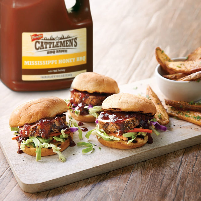 ["Three mini burgers with coleslaw and Cattlemen's Mississippi Honey BBQ sauce on a wooden cutting board with potato wedges in the background."]
