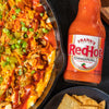 A cast iron skillet filled with a cheesy dip topped with Franks RedHot Original Cayenne Pepper Sauce green onions and tortilla chips.