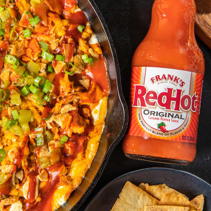 A cast iron skillet filled with a cheesy dip topped with Franks RedHot Original Cayenne Pepper Sauce green onions and tortilla chips.
