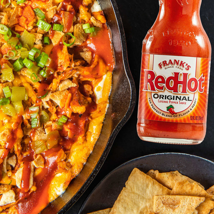 A cast iron skillet filled with a cheesy dip topped with Frank's RedHot Original Cayenne Pepper Sauce, green onions, and tortilla chips.