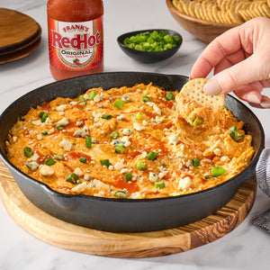 A cast iron skillet filled with buffalo chicken dip topped with crumbled blue cheese, green onions, and Franks RedHot sauce.