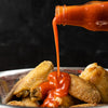 A bottle of hot sauce is pouring sauce over fried chicken wings in a bowl.