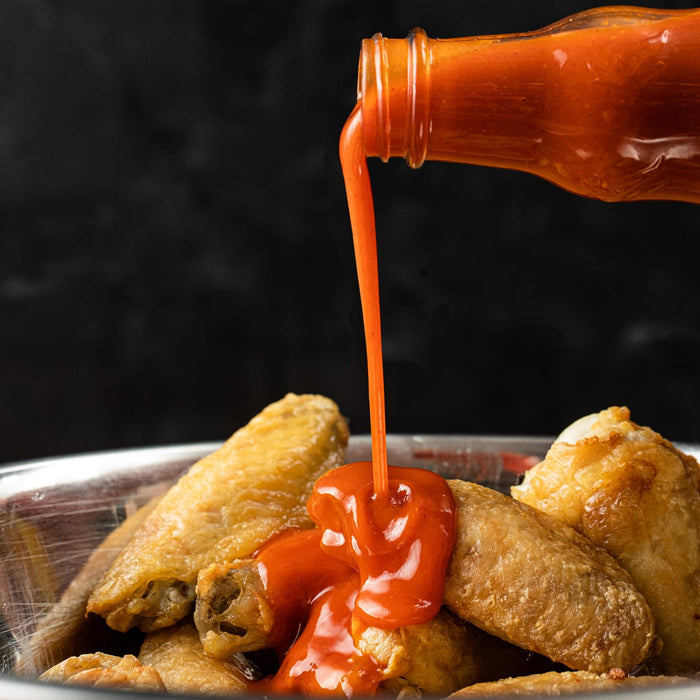 A bottle of hot sauce is pouring sauce over fried chicken wings in a bowl.