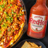 A cast iron skillet filled with a cheesy, chicken, and buffalo wing dip with a bottle of Frank's RedHot Original Cayenne Pepper Sauce.