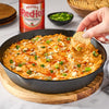 A cast iron skillet filled with buffalo chicken dip topped with crumbled blue cheese and green onions, with a hand dipping a cracker into the dip, and a bottle of Frank's RedHot Original Cayenne Pepper Sauce in the background.