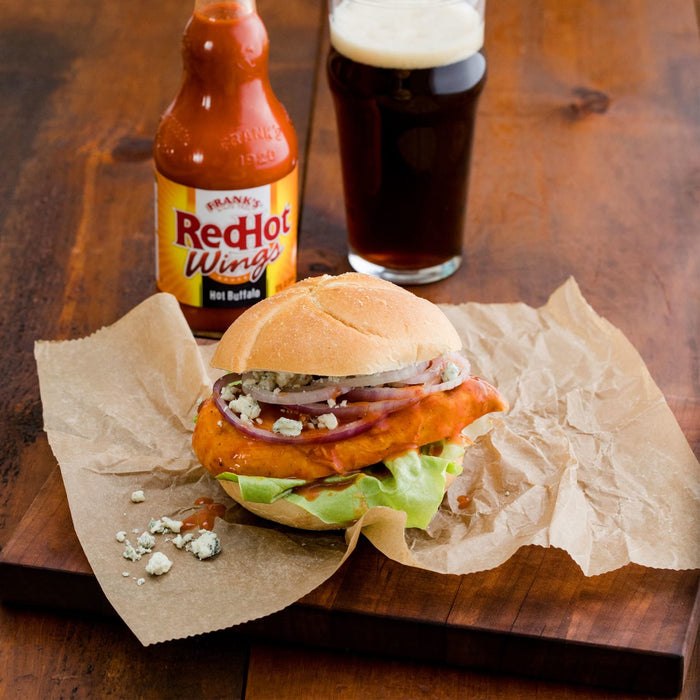 ["A Frank's RedHot Wings sandwich with blue cheese and red onion on a bun, next to a glass of beer and a bottle of Frank's RedHot."]