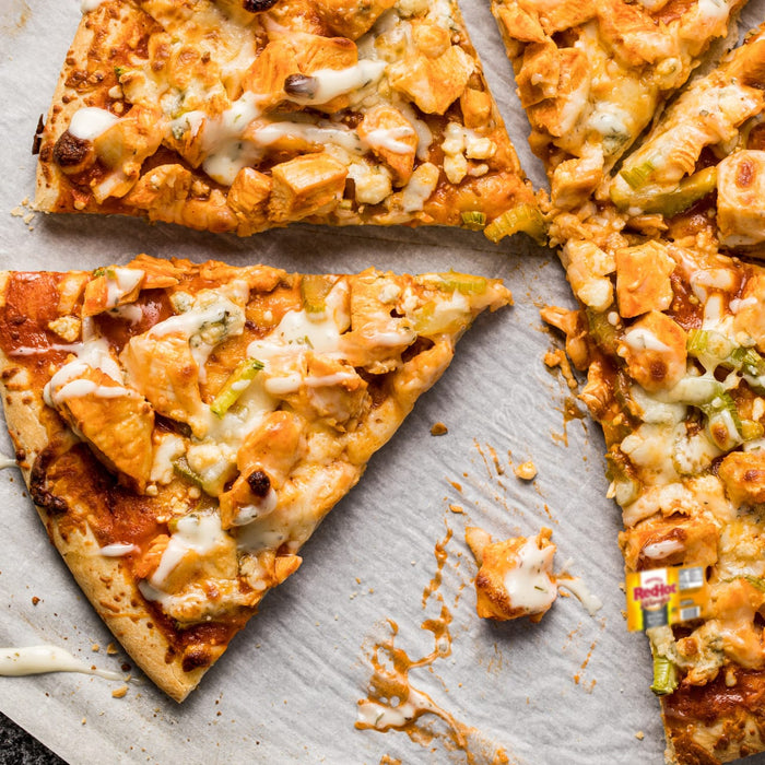 A man in a white hat and gloves cuts a pizza with a pizza cutter in a kitchen.