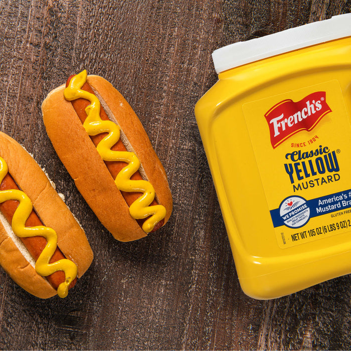 A jar of Frenchs Classic Yellow Mustard next to two hot dogs with mustard.