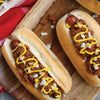 Two hot dogs with chili, mustard and onions on a wooden cutting board.