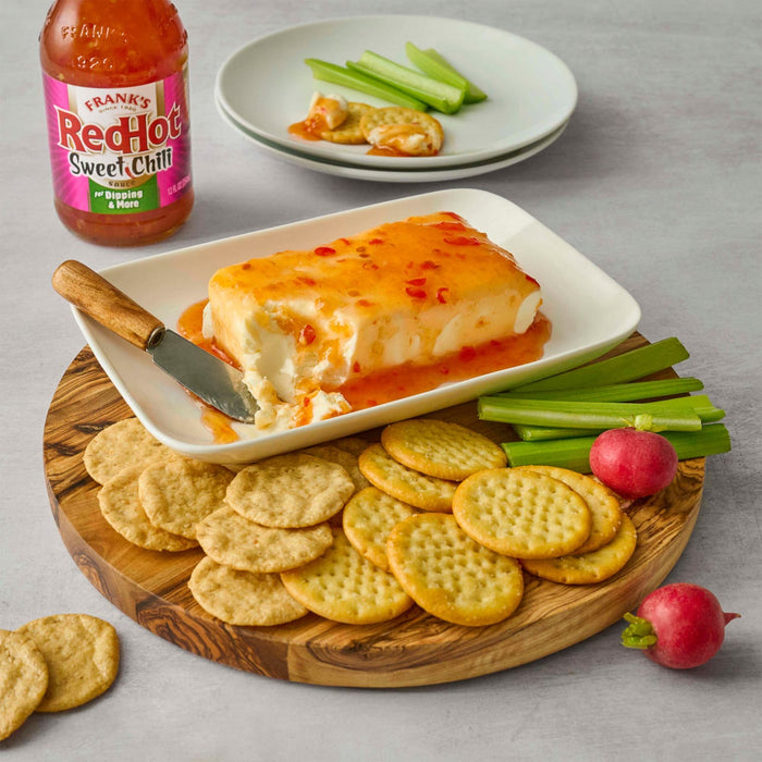 A block of cream cheese topped with Frank's RedHot Sweet Chili sauce is served with crackers, celery, and radishes.