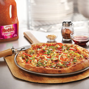 A Frank's RedHot Sweet Chili pizza sits on a wooden peel with a pizza cutter, alongside a bottle of Frank's RedHot Sweet Chili sauce, crushed red pepper flakes, and a small bowl of sauce.
