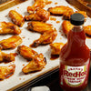 A tray of glazed chicken wings sits next to a bottle of Franks Red Hot Stingin Honey Teriyaki Wing Hot Sauce Mild 12 Oz.