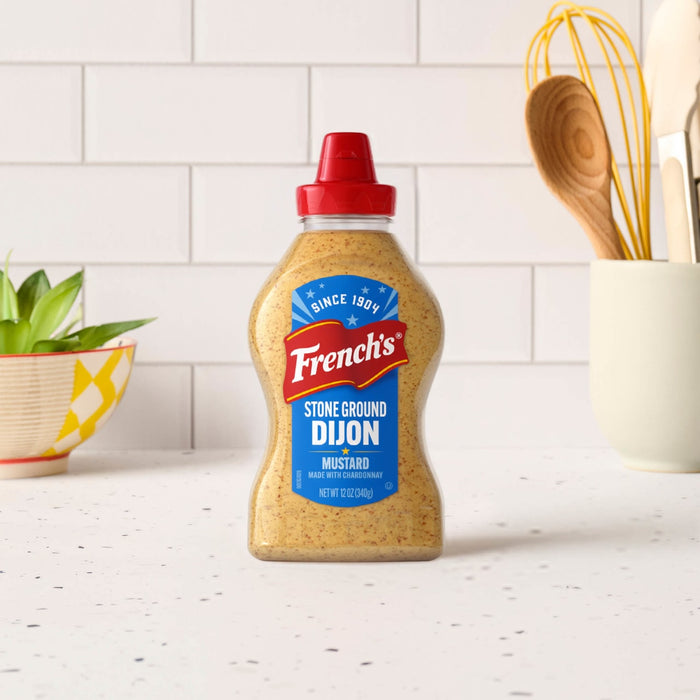 A bottle of Frenchs Beige Stone Ground Dijon Mustard 12 Oz sits on a white speckled countertop in a bright kitchen setting with cooking utensils in the background.