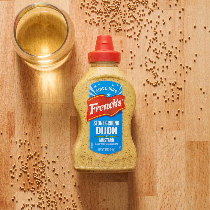 A bottle of Frenchs Beige Stone Ground Dijon Mustard 12 Oz sits on a wooden surface surrounded by scattered mustard seeds and a glass of white wine.