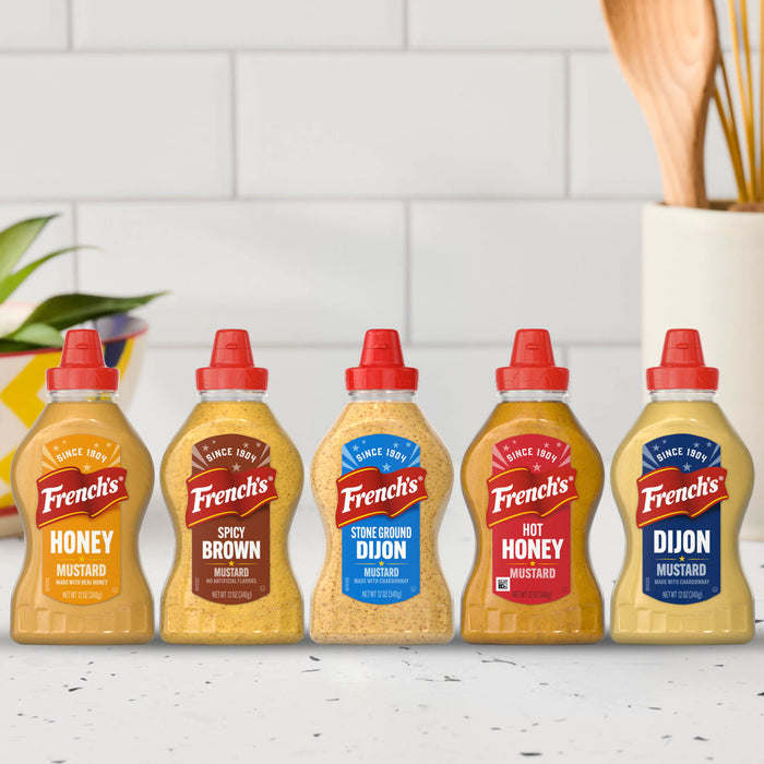 A lineup of five Frenchs mustard varieties including the Frenchs Beige Stone Ground Dijon Mustard 12 Oz displayed against a white tiled kitchen background.