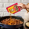 A hand pours a packet of Cholula Medium Taco Seasoning Mix 1 Oz over browned ground meat in a blue skillet to add authentic flavor.