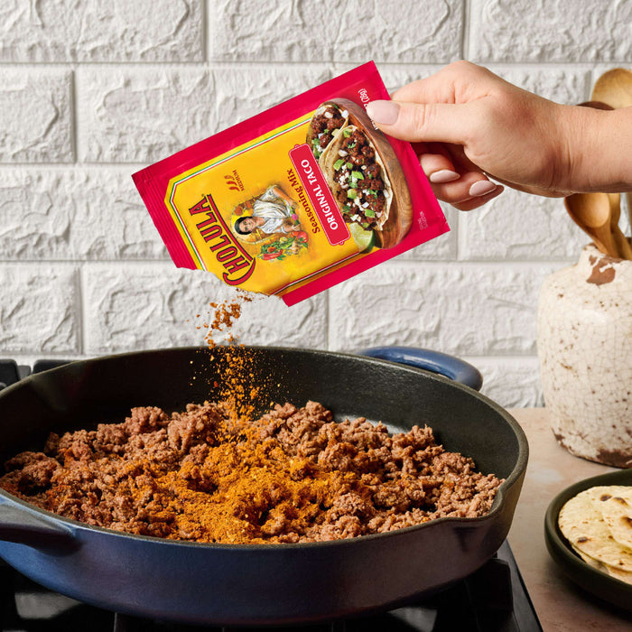 A hand pours a packet of Cholula Medium Taco Seasoning Mix 1 Oz over browned ground meat in a blue skillet to add authentic flavor.