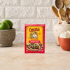 A packet of Cholula Medium Taco Seasoning Mix 1 Oz stands on a kitchen counter surrounded by fresh limes, garlic, and wooden cooking utensils.