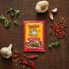 A packet of Cholula Medium Taco Seasoning Mix 1 Oz is displayed on a dark wooden surface surrounded by fresh garlic bulbs, sprigs of oregano, and dried red chili peppers.
