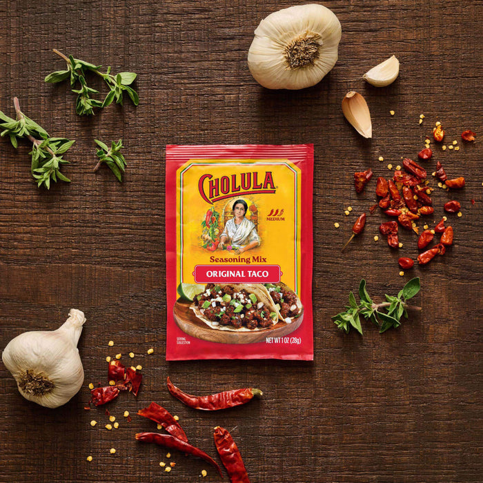 A packet of Cholula Medium Taco Seasoning Mix 1 Oz is displayed on a dark wooden surface surrounded by fresh garlic bulbs, sprigs of oregano, and dried red chili peppers.