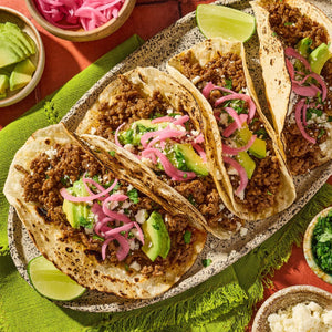 A platter of three street tacos filled with ground beef seasoned with Cholula Medium Taco Seasoning Mix 1 Oz and topped with avocado slices and pickled red onions.